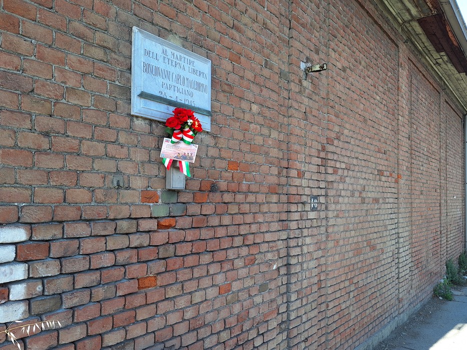 Lapide commemorativa su muro di mattoni, adornata con una corona di fiori e un nastro tricolore.