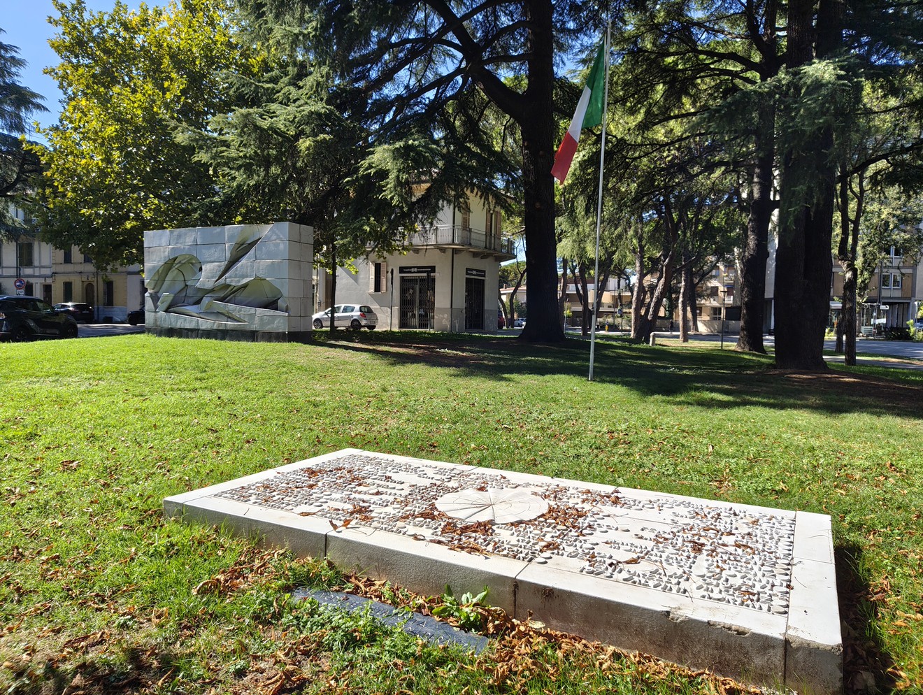 Lastra orizzontale con iscrizione su prato, monumento scultoreo in pietra e alberi sullo sfondo