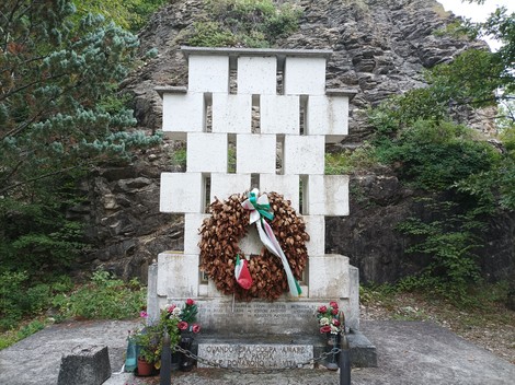 Monumento ai caduti di Rio Farnese, con una corona di fiori e decorazioni, circondato da natura.