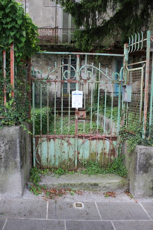 Porta arrugginita con cancello verde e vegetazione che la circonda, rappresenta una pietra d'inciampo a Sirtoli.