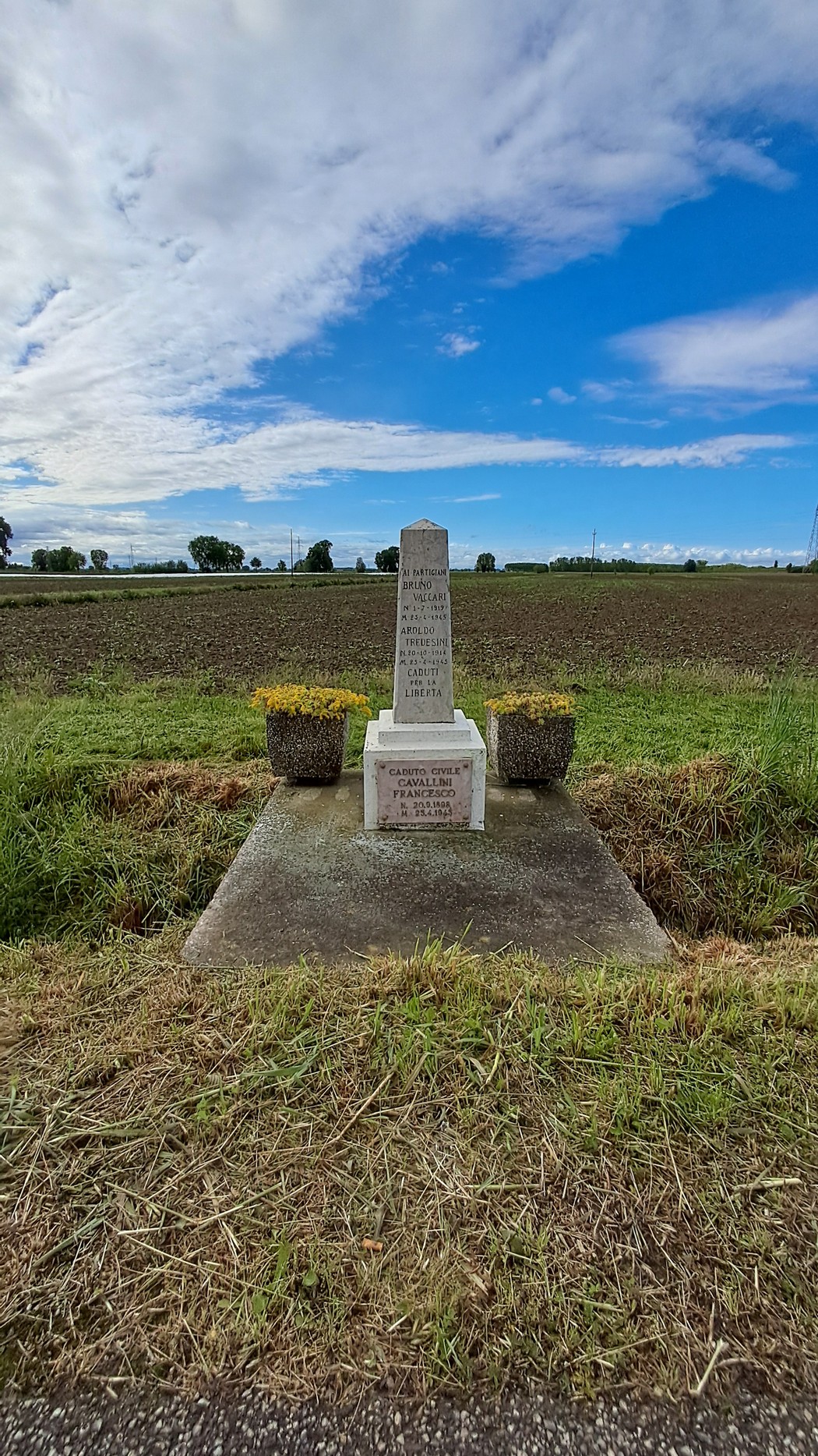 Cippo commemorativo incastonato in un paesaggio rurale, circondato da fiori e cielo blu.