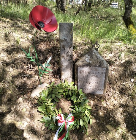 Cippo commemorativo di Uscieri con un omaggio floreale e una corona di alloro in un contesto naturale.