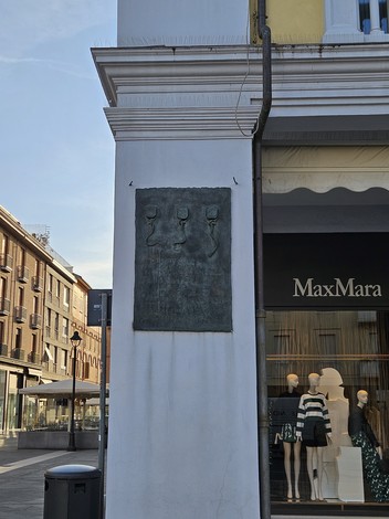 Targa commemorativa ai Tre Martiri, con rilievi su fondo scuro, affissa a un muro bianco in un'area urbana.