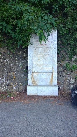 Stele commemorativa dedicata alle Brigate Partigiane, immersa nella natura, con un muro di pietra sullo sfondo.
