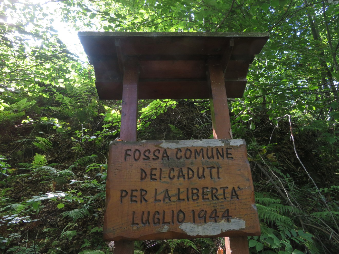 Una targa in legno indica "Fossa comune dei caduti per la libertà" circondata da vegetazione.