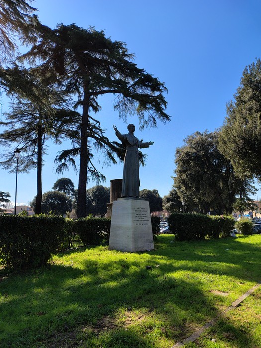 Statua di Pacelli circondata da alberi in un parco soleggiato. Fondo verde con cielo blu sereno.