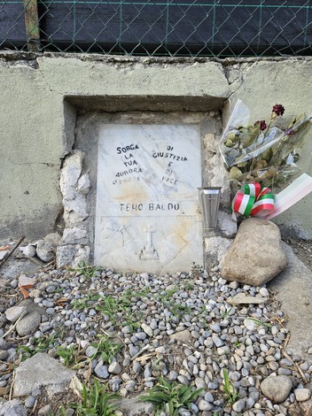 Cippo commemorativo con fiori e una candela, situato in un'area con ciottoli e vegetazione.