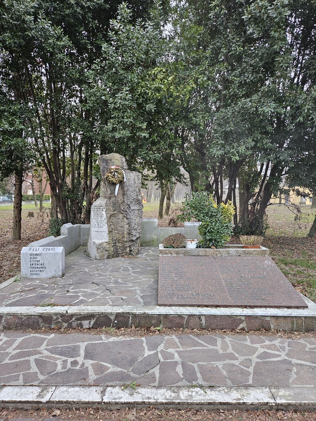 Monumento ai Caduti con una stele di pietra circondata da piante e fiori, immerso nel verde.