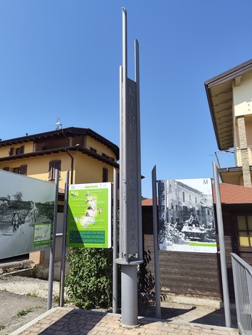 Pannelli informativi e un totem metallico con scritta verticale, tra cui foto storiche in bianco e nero, in un'area urbana.
