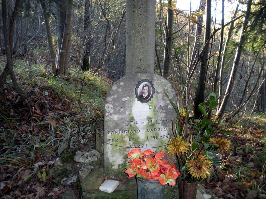 Cippo commemorativo di un partigiano caduto, adornato con fiori, immerso nella natura boschiva.
