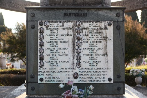 Sacrario ai caduti con nomi e date in memoria dei partigiani, circondato da fiori e natura.