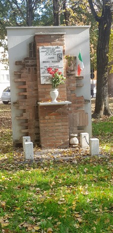 Monumento a Rizzieri, con fiori e bandiera italiana, circondato da alberi e foglie.