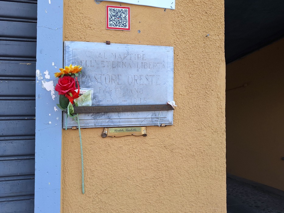 Lapide in marmo con iscrizione su parete esterna gialla, ornata da una rosa e un fiore finti.