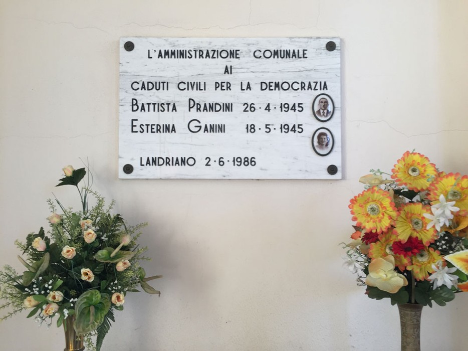 Lapide commemorativa ai caduti civili per la democrazia, con nomi e date, decorata con fiori.