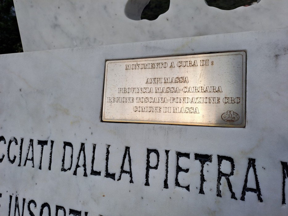 Targa metallica con iscrizione fissata su monumento in marmo bianco, sopra scritte incise nella pietra.