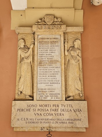 Lapide commemorativa con iscrizioni in onore dei caduti per la libertà, adornata da figure simboliche.