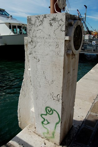 Cippo commemorativo con iscrizioni e un graffito, situato vicino al mare, dedicato ai dipendenti Acnil caduti.