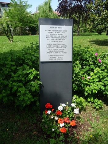 Stele commemorativa al Campo PG 62 della Grumellina, circondata da vegetazione e fiori colorati.