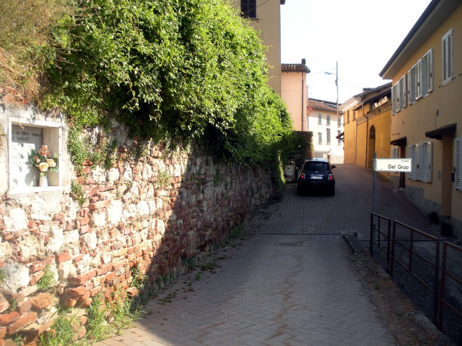 Strada acciottolata con muratura in pietra e vegetazione, presenta una lapide e una macchina parcheggiata.