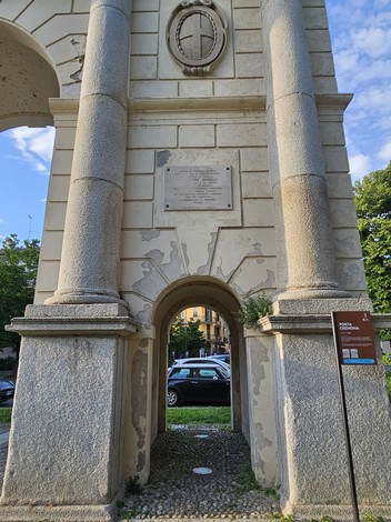 Lapide commemorativa incastonata in un arco architettonico con colonne, circondata da vegetazione e auto parcheggiate.