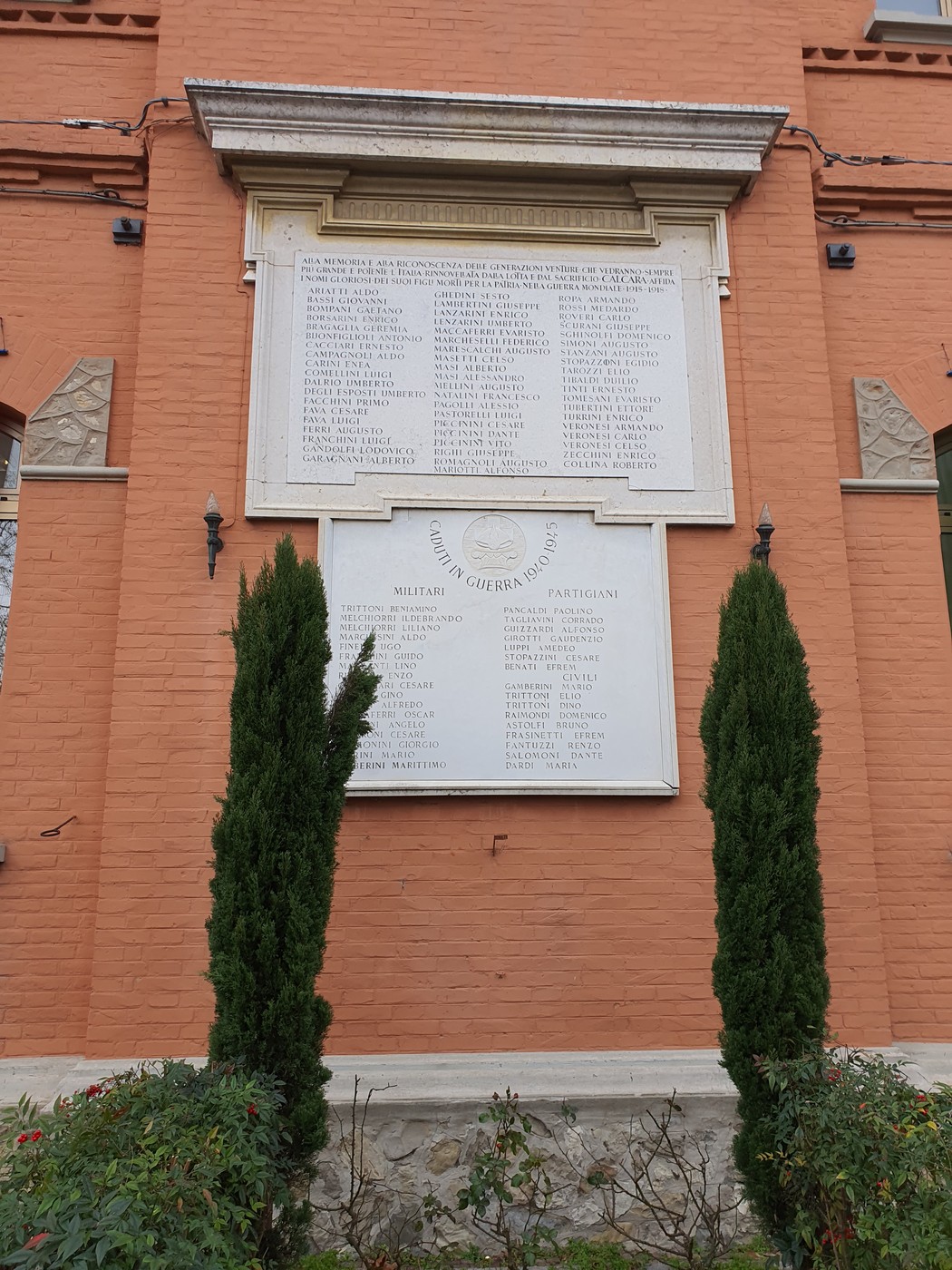 Lapide commemorativa ai caduti, con nomi incisi, situata su una parete di mattoni rossi, circondata da piante.