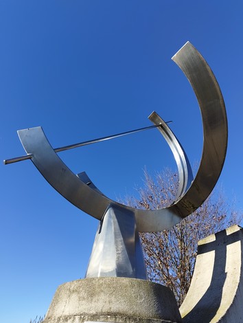 Monumento commemorativo in metallo lucido con forme moderne, contro un cielo blu sereno.