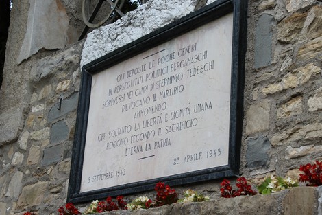 Lapide commemorativa dedicata ai perseguitati politici, incastonata in un muro di pietra con fiori rossi.