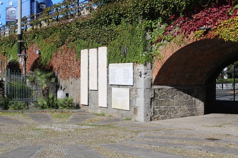 Lapide commemorativa circondata da vegetazione, antica muratura e architettura storica.