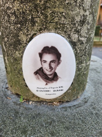 Cippo commemorativo con targa in ceramica ritraente Giovanni Oldani, situato su un supporto di pietra.
