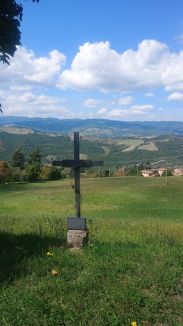 Vista panoramica con croce in legno su un prato verde, circondata da colline e un cielo blu con nuvole bianche.