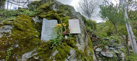 Lapide commemorativa con fiori, situata su una roccia circondata da natura.