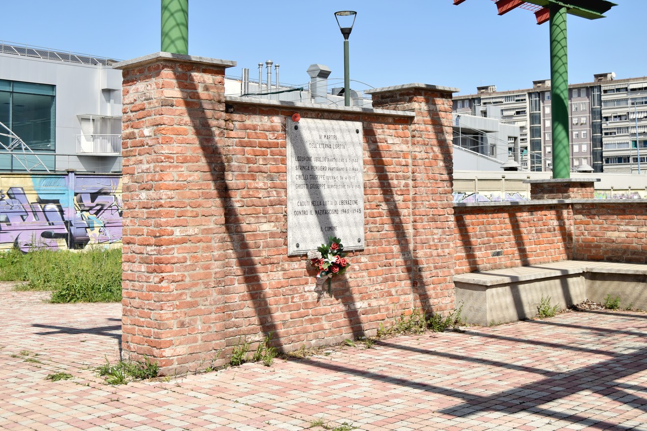 Monumento commemorativo con una parete di mattoni, decorato con fiori, in un'area urbana aperta.
