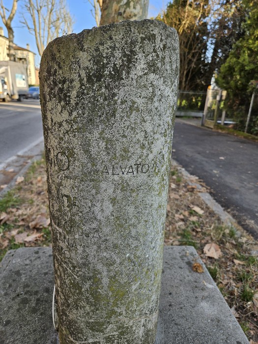 Cippo in pietra con iscrizioni visibili, circondato da una strada e vegetazione.