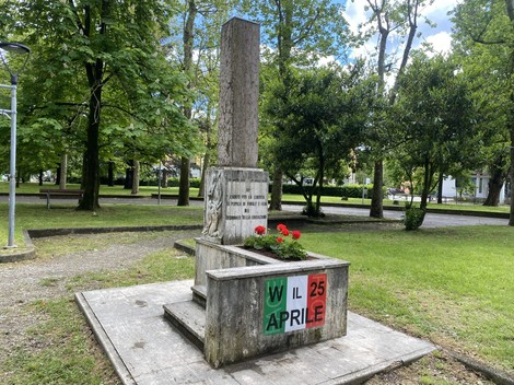 Monumento ai caduti, con iscrizioni e fiori rossi, immerso in un parco verde.