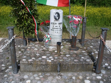 Cippo commemorativo di Azzolini, decorato con fiori e bandiere, in un contesto naturale di verde e fiori.