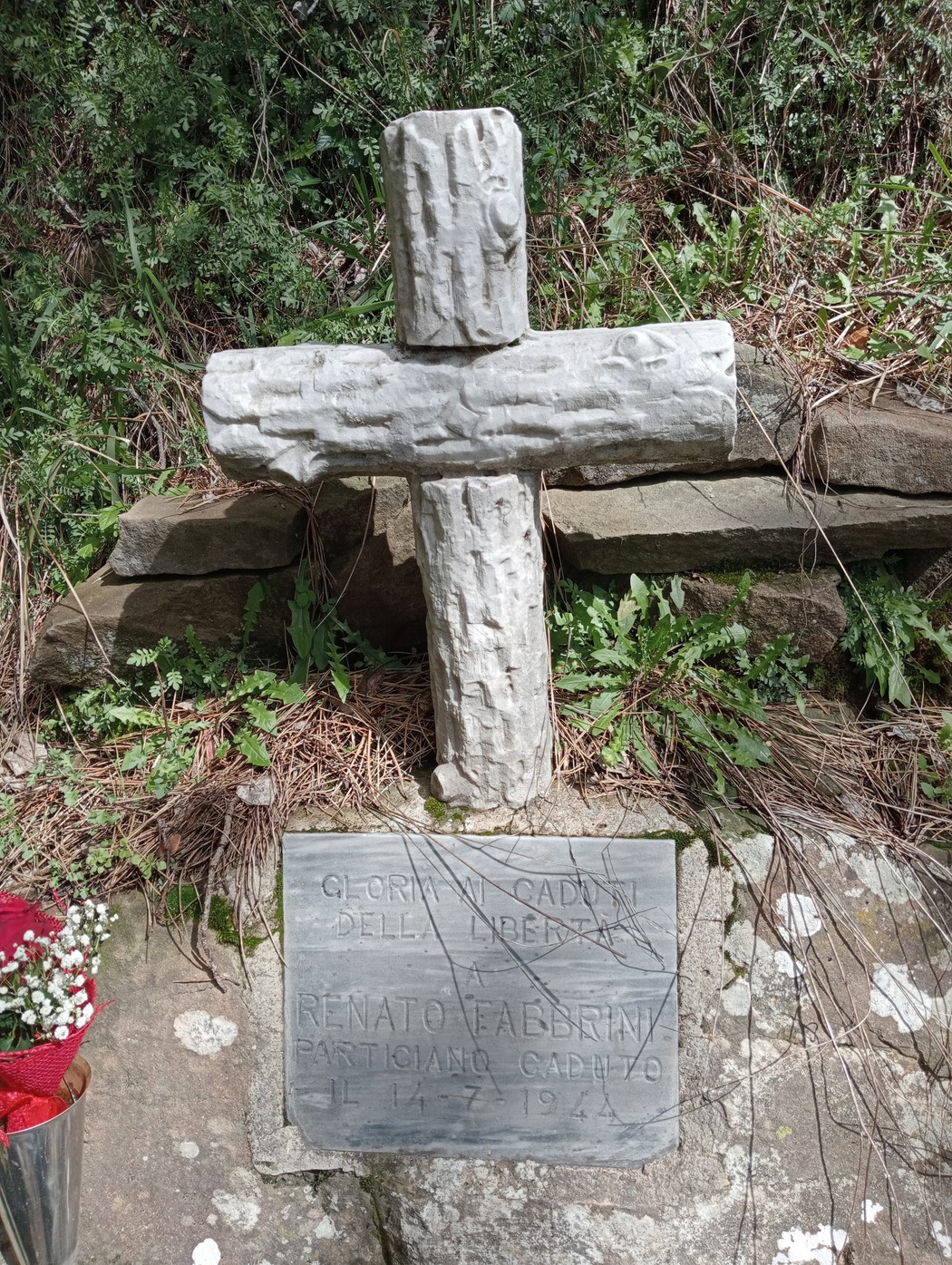 Cippo commemorativo con croce in legno e lapide dedicata a Renato Fabbrini, in un ambiente naturale.