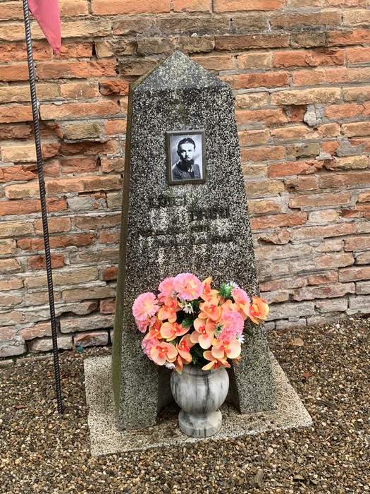 Cippo commemorativo con fotografia e fiori, situato in un contesto storico.