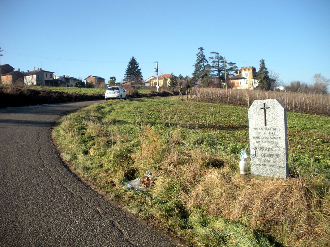 Cippo commemorativo lungo una strada rurale, circondato da un paesaggio verde con case sullo sfondo.