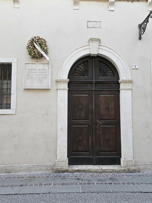 Lapide commemorativa su una parete accanto a una porta in legno, decorata con una corona di fiori.