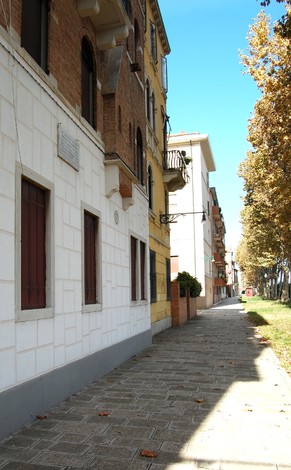 Vista di una strada con edifici storici e foglie che cadono dagli alberi ai lati. Atmosfera tranquilla e soleggiata.