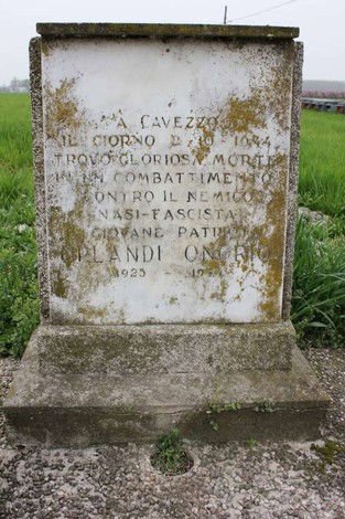 Cippo commemorativo in pietra con iscrizioni, immerso in un paesaggio verde di campagna.