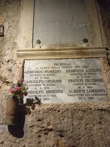 Lapide in marmo con iscrizione, fissata a una parete in pietra, con un piccolo vaso di fiori a lato.