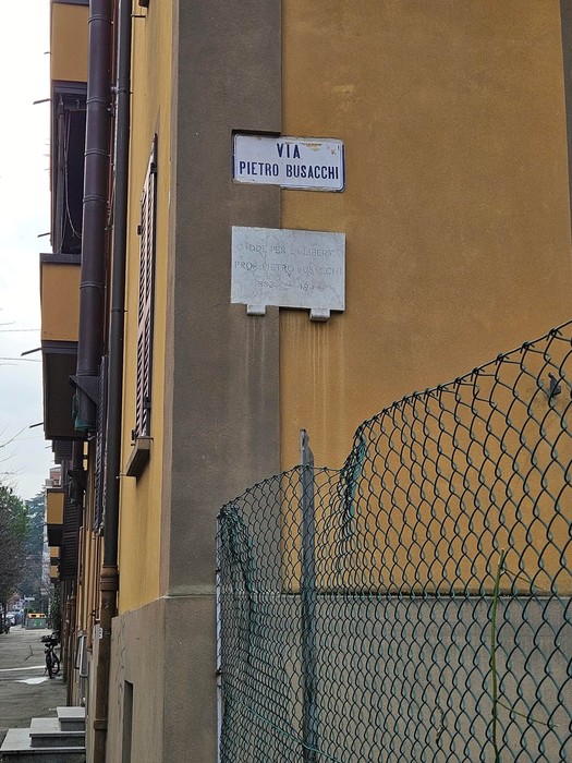 Lapide commemorativa su un muro angolare di un edificio, con insegna che indica via Pietro Busacchi.
