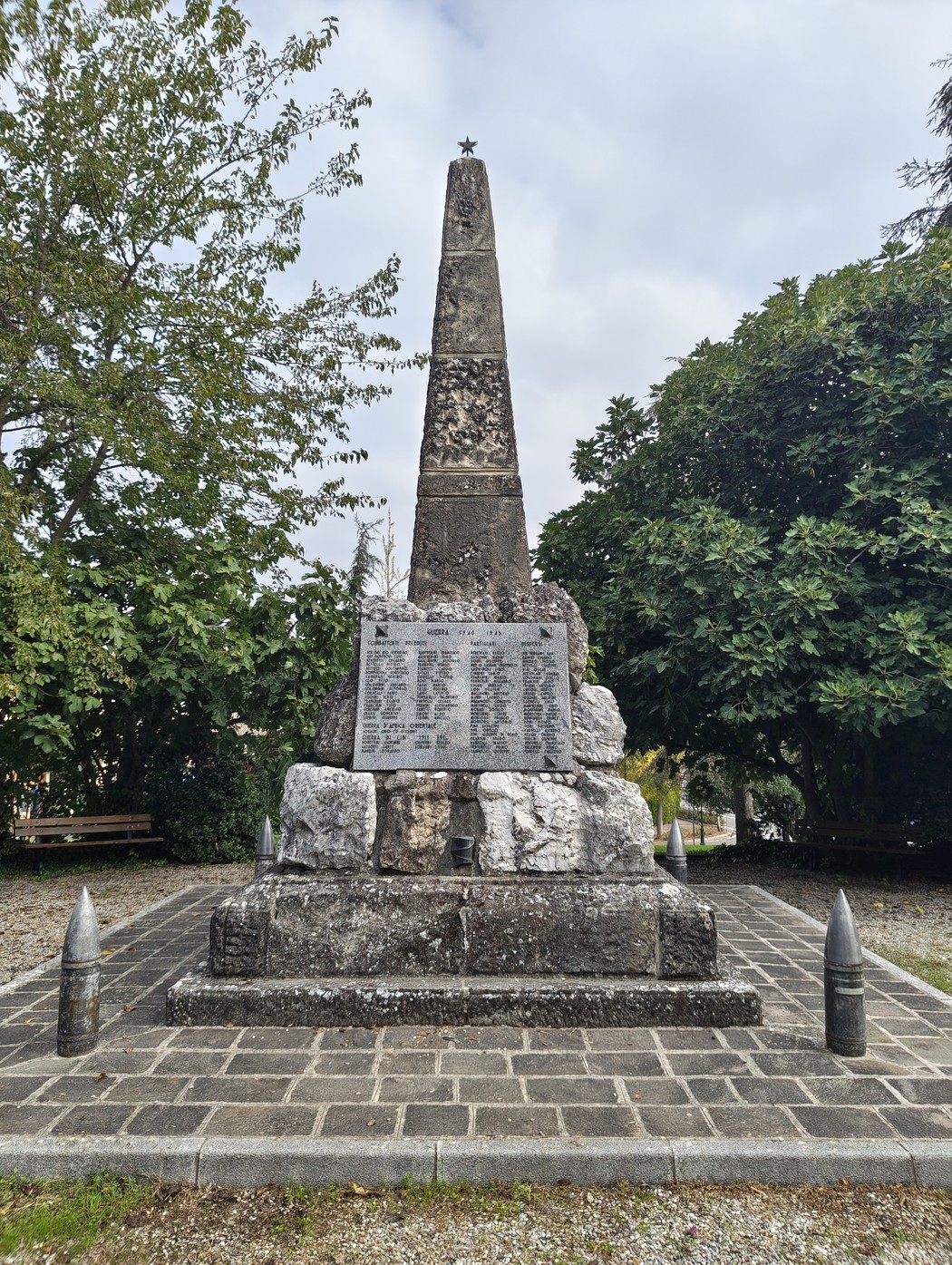 Monumento a obelisco in pietra con lapide incisa, su basamento roccioso in uno spiazzo alberato.