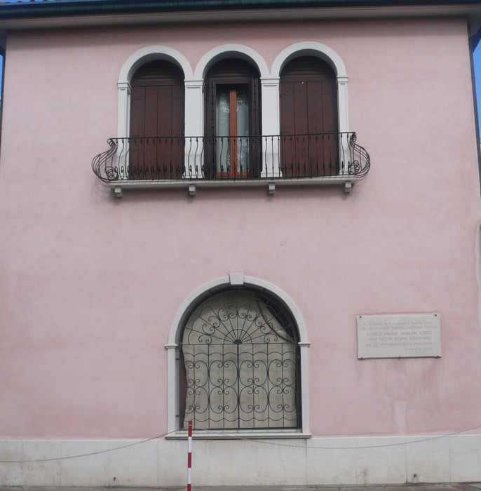 Lapide commemorativa su una facciata di un edificio rosa con balcone e decorazioni in ferro battuto.