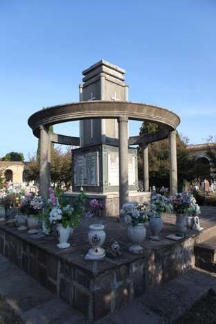 Sacrario ai caduti con monumento centrale, circondato da fiori e vasi in un ambiente sereno.
