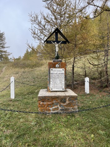 Monumento commemorativo con croce nera su base in pietra immerso nella natura, circondato da alberi.