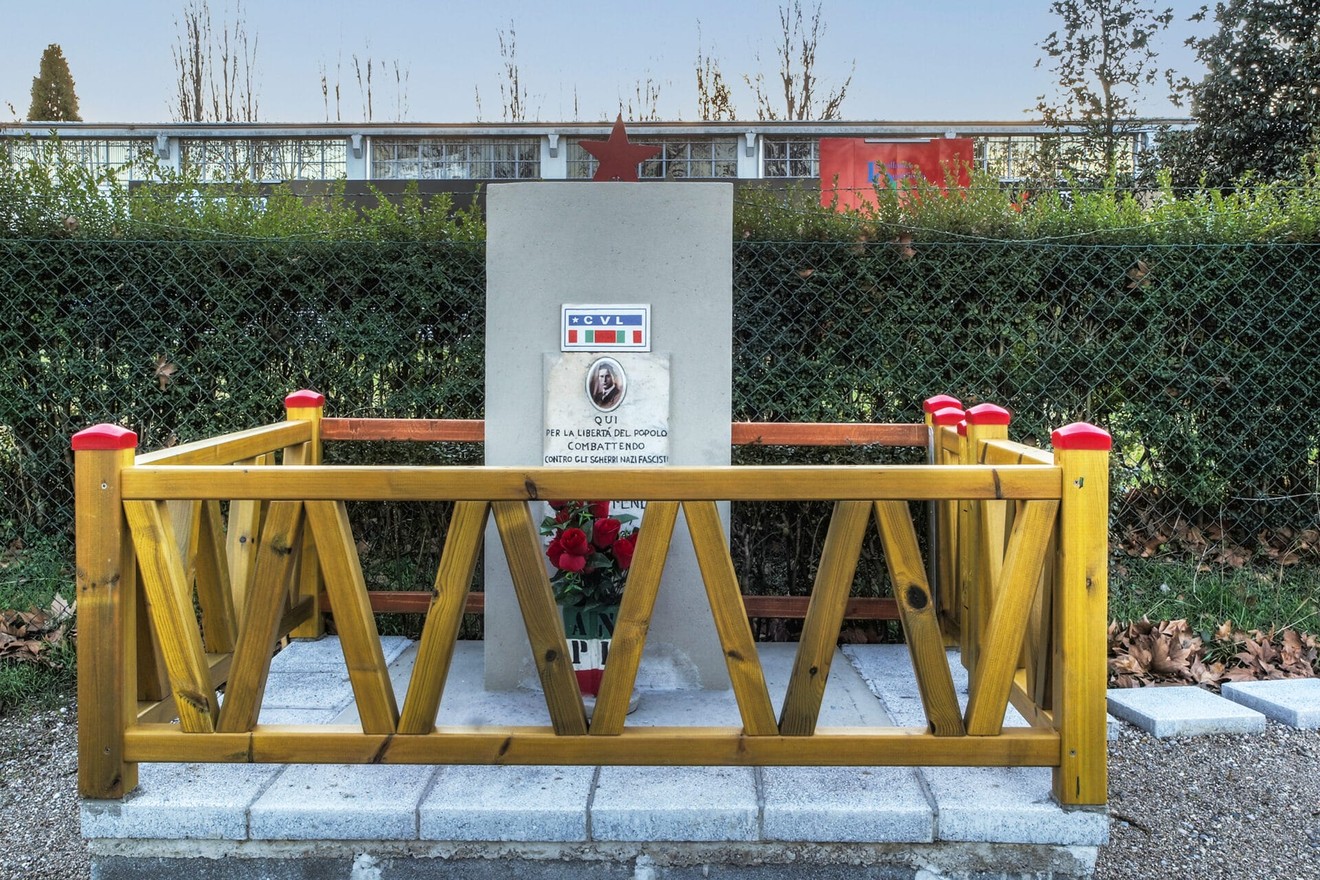 Cippo commemorativo dedicato a Benetti, circondato da una recinzione in legno e fiori.