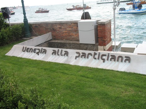 Monumento alla partigiana affacciato sull'acqua, con imbarcazioni sullo sfondo e un'area verde circostante.