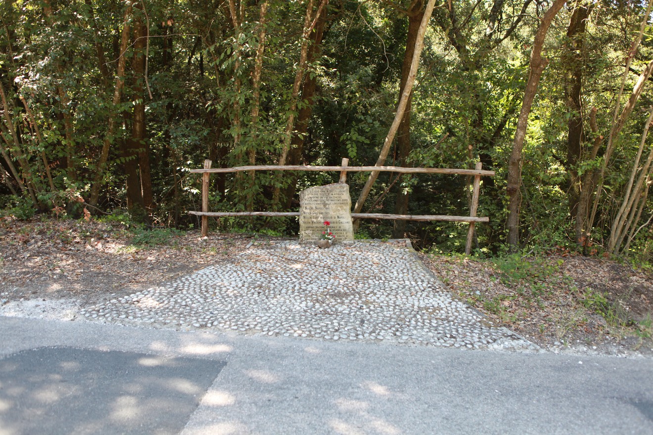 Cippo dedicato alla famiglia Nocchi, immerso nel verde e circondato da alberi, con selciato acciottolato.